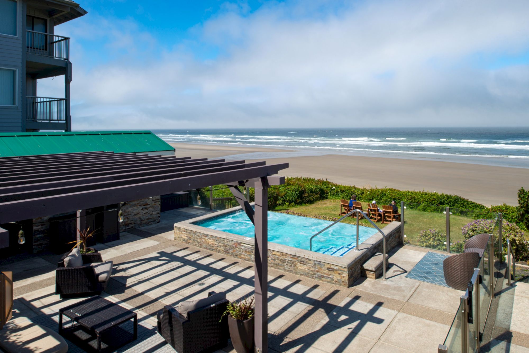 A sunny beach house with a shaded wooden patio, a small pool, lounge chairs, and a wide sandy shore beyond the ocean under blue skies.