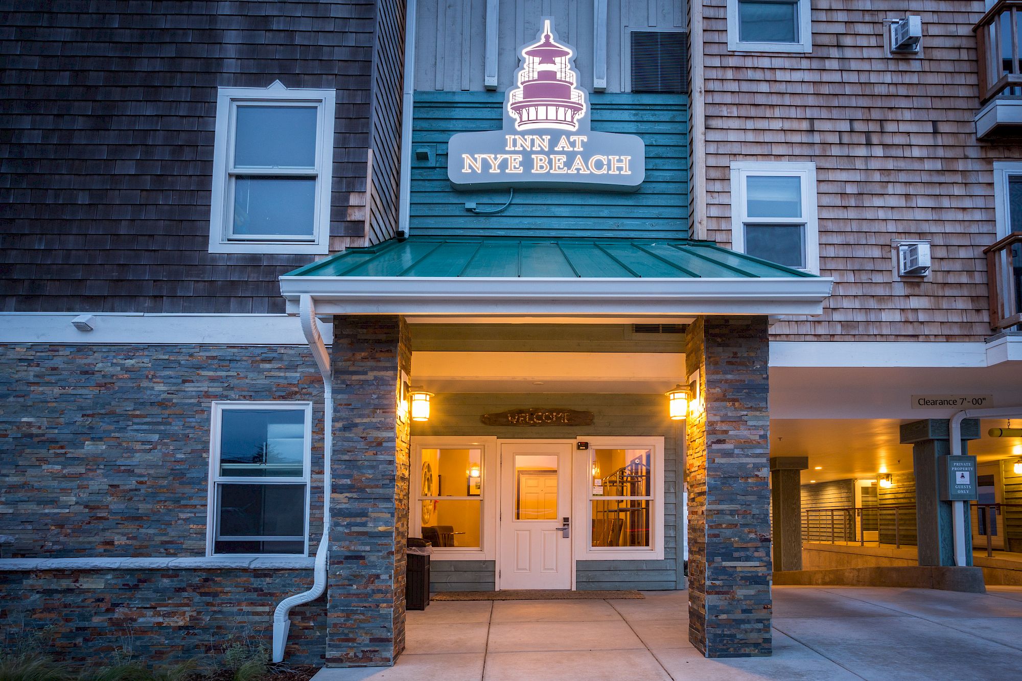 A New England-style inn or hotel entrance with a sign reading &ldquo;New Inn at NYJB&hellip; Block&rdquo; above a lit doorway under a green roof, brick and stone exterior.