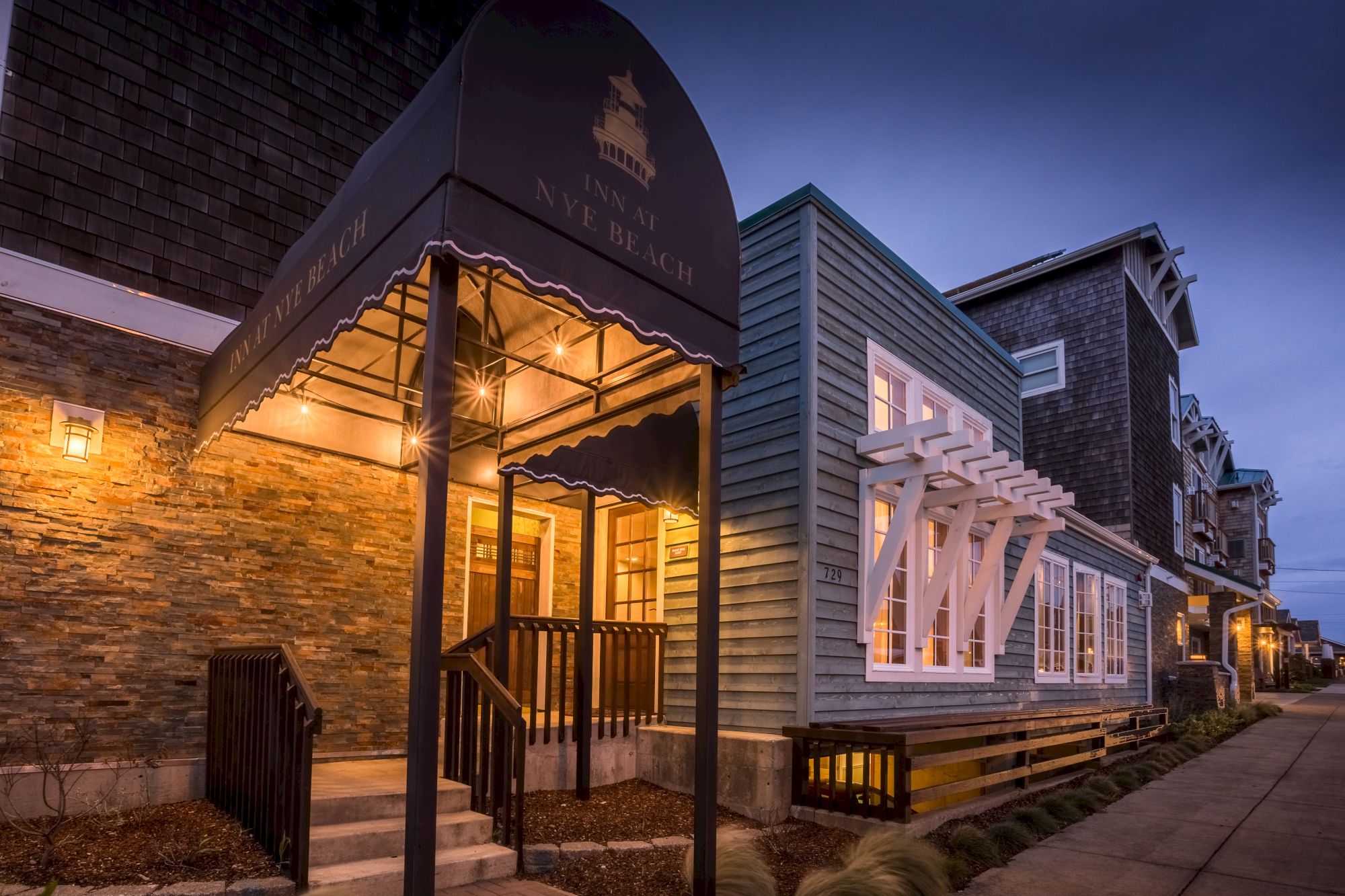 A lit storefront with a curved awning and white window frames on a brick-and-siding exterior along a quiet street at dusk.