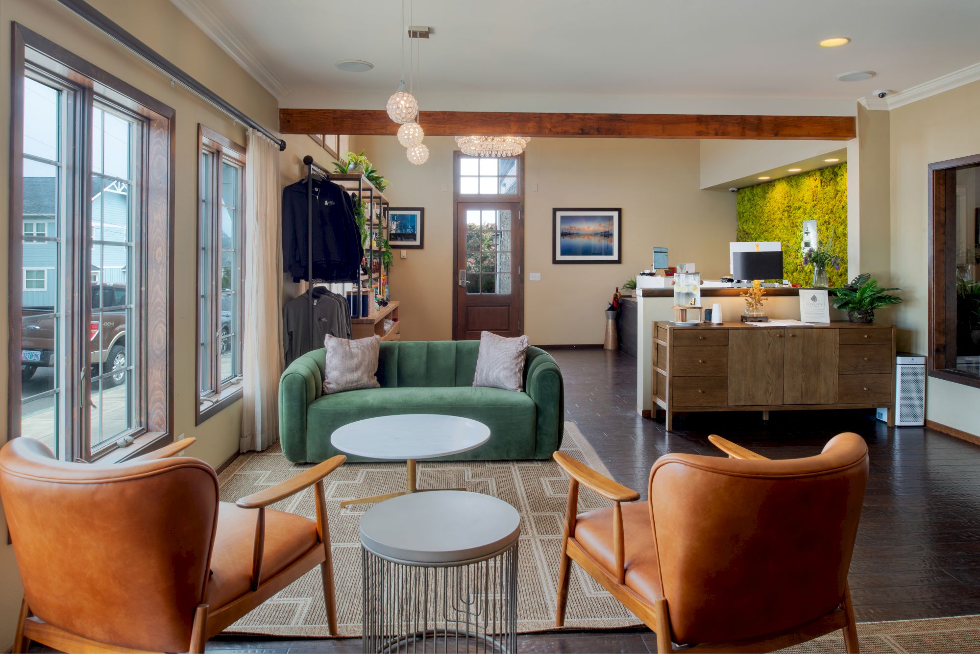 A modern, cozy showroom or lobby with a green sofa, two tan leather chairs, white round tables, and a small metal side table, near large windows and a reception desk.