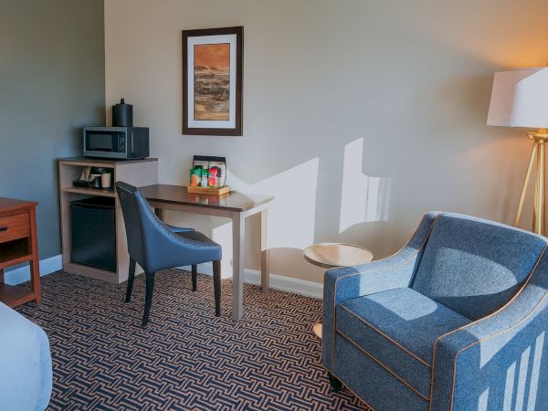A hotel room with a desk, chair, mini-fridge, microwave, framed art, a blue armchair, a floor lamp, and patterned carpet, light streaming through.