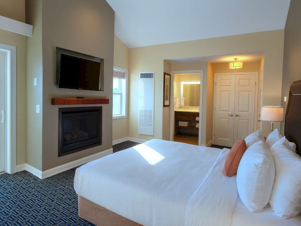 A bright hotel room with a king bed, white sheets, multiple pillows, a mounted TV above a fireplace, and a doorway leading to a bath and hallway.