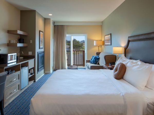 A hotel room with two queen beds, a sitting area, kitchenette, desk, and balcony overlooking trees; warm lighting and neutral decor.
