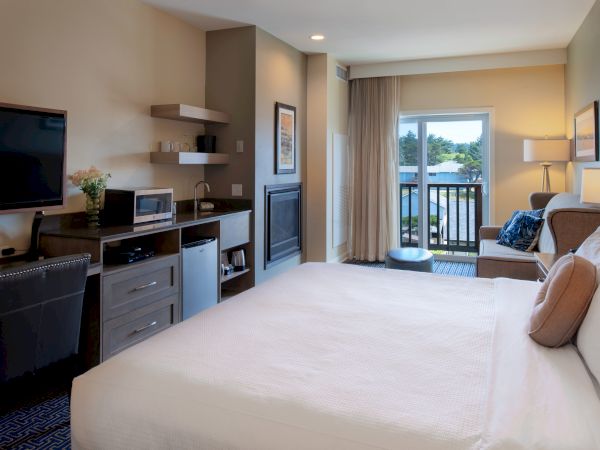A modern hotel room with a king bed, a desk area, a kitchenette, fireplace, and a balcony overlooking trees.