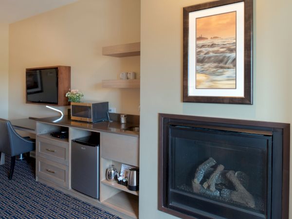A hotel room area with a desk, mini-fridge, microwave, and coffee setup, plus a framed artwork and a gas fireplace; the carpet is patterned.