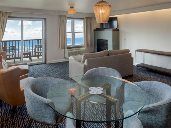 A modern hotel suite with a glass dining table for four, gray couches, a fireplace, and a balcony overlooking the ocean under soft lighting.
