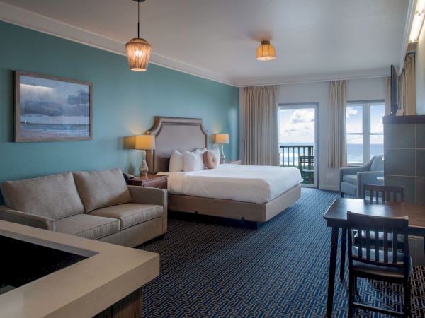 A bright hotel room with a teal accent wall, king bed, two bedside lamps, a gray sofa, dark wood desk and chairs, and a balcony view, cozy and modern.