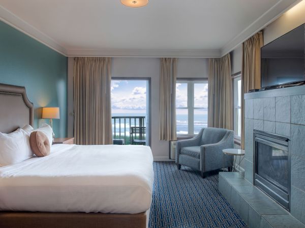 A hotel room with a king bed, teal accent wall, bedside lamps, a seating area, fireplace, and a balcony overlooking water. Ends here.