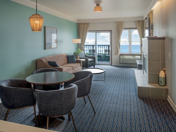 Cozy hotel living area with teal accent wall, round dining table and four chairs, beige sofa, fireplace, large windows, and balcony view.