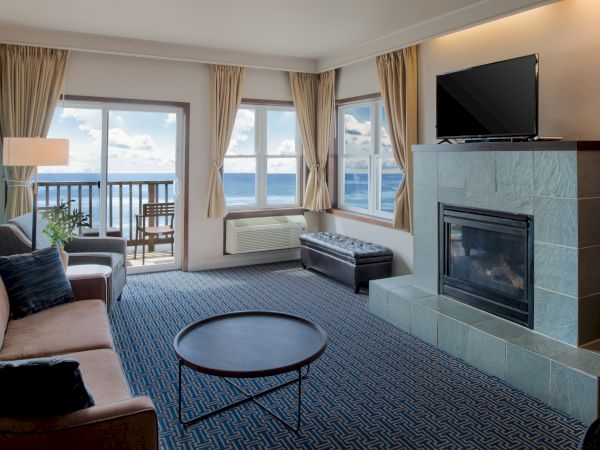 A cozy hotel living room with a blue carpet, beige sofa, round coffee table, and a fireplace; large windows open to a balcony with ocean views.