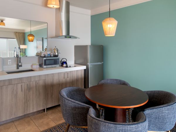 A compact kitchen and dining nook with a round black table, four grey chairs, a teal accent wall, and a small kitchenette with wooden cabinets and a fridge, all under pendant lighting.