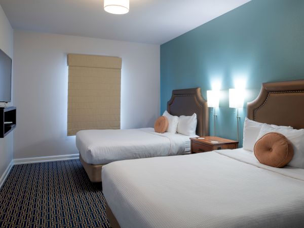 A hotel room with two queen beds, teal accent wall, nightstands, round pillows, a wall-mounted TV, window with shade, and soft overhead lighting. End.
