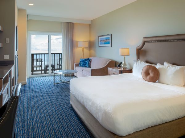 A bright hotel room with a king bed, seating area, desk, balcony view, and soft blue-gray decor, ending with a neat, inviting vibe.