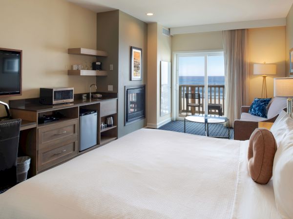 A hotel room with a bed in foreground, a kitchenette/desk area on the left, a fireplace, and a balcony with a view outside.