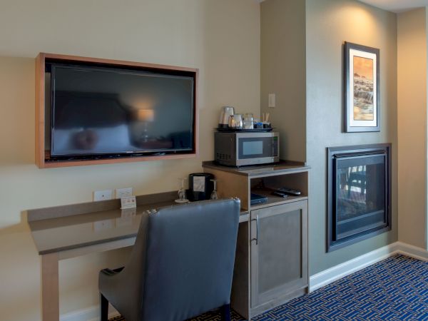 A small hotel-style workspace with a wall-mounted TV, a desk and chair, a microwave on a cabinet, a counter, and a gas fireplace in a cozy suite.