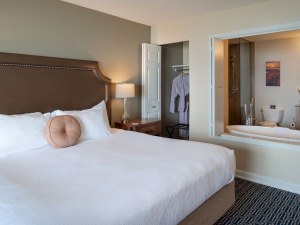 A hotel room with a large bed, nightstands, a wardrobe, a mirror, and an open bathroom area with a tub. 140 characters ending with a period.