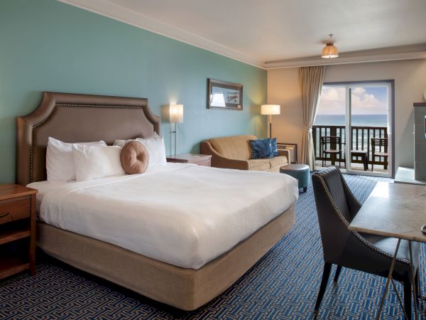 A hotel room with a king bed, teal walls, two nightstands, a sofa, desk chair, and a balcony overlooking the ocean.