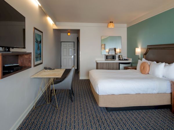 A hotel room with a queen bed, teal accent wall, desk and chair, wall-mounted TV, dresser with mirror, and a small table setup near the entry.