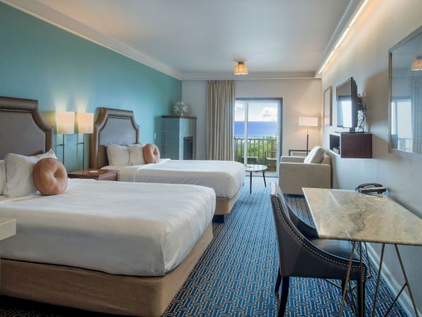 A hotel room with two queen beds, teal accent wall, desk, chairs, and a balcony view; bright, modern and inviting.