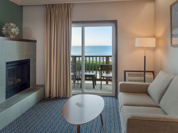 A cozy living room with a fireplace, blue patterned rug, beige sofa, and a balcony view of the sea through glass doors.