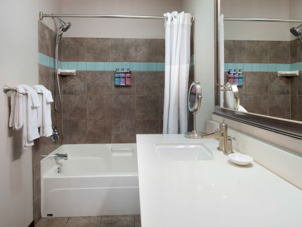 Modern bathroom with a white bathtub/shower combo, gray tile walls, a long white counter, and a large mirror; towels and toiletries are neatly arranged.
