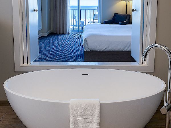 A modern hotel bathroom with a freestanding oval bathtub and a towel draped over the edge, framed by a large window looking into a bedroom with a bed and balcony view.