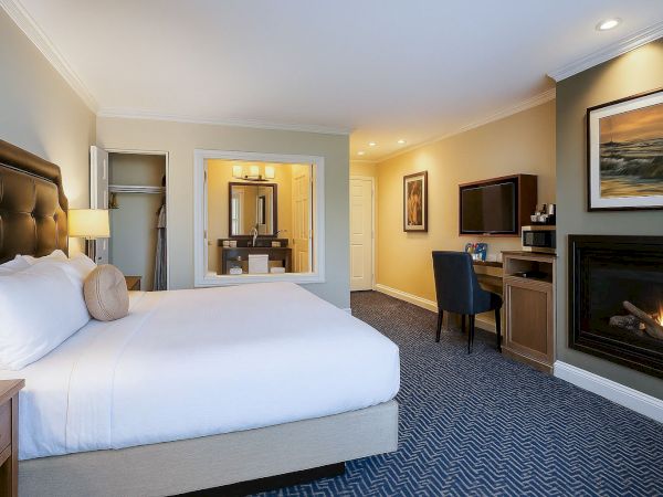 A hotel room with a king bed, a workspace with desk and chair, a fireplace, wall-mounted TV, and a seating area; warm lighting and a neutral decor.