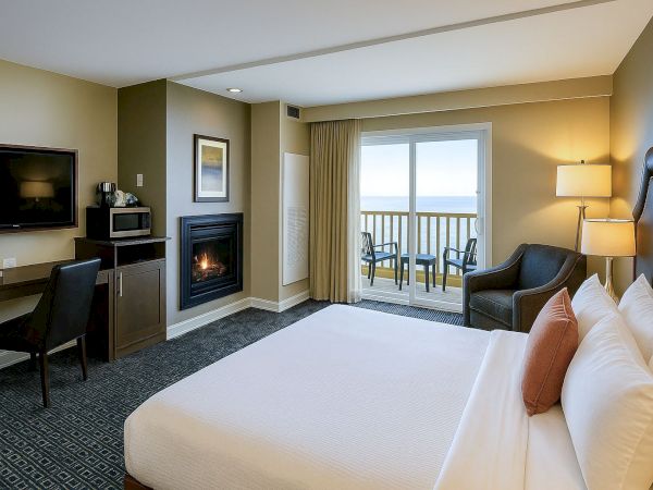 A modern hotel room with a king bed, fireplace, desk, flat-screen, and a balcony overlooking water; cozy seating and warm lighting.