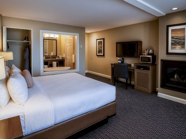 A tidy hotel room with a large bed, nightstands, a sofa chair, a desk, a mounted TV, a fireplace, and an open doorway to a bathroom, cozy lighting.