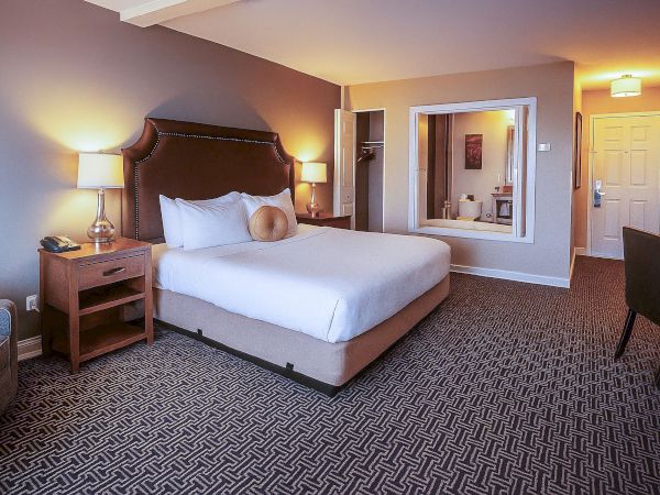 A hotel room with a large bed, nightstands, lamps, a seating area, and a view into a bathroom or vanity area; warm lighting throughout.