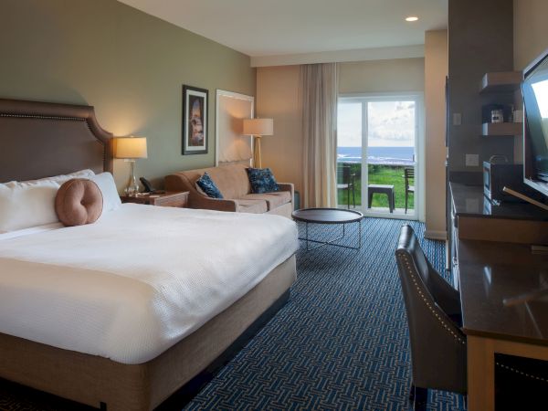 A hotel room with a large bed, seating area, desk, and balcony overlooking water; modern decor and warm lighting. End with a period.