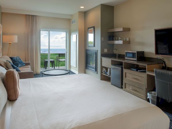 A cozy hotel suite with a king bed, sitting area, desk, fireplace, and a balcony view of the ocean, bright natural light, and soft decor.
