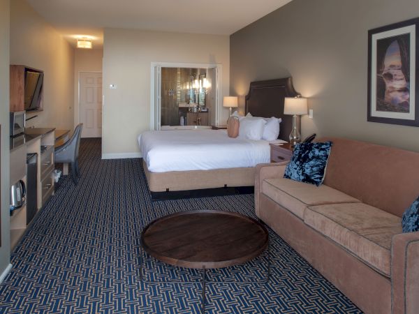 A hotel room with a bed, sofa, desk, round coffee table, and a large mirror; blue patterned carpet and framed wall art.