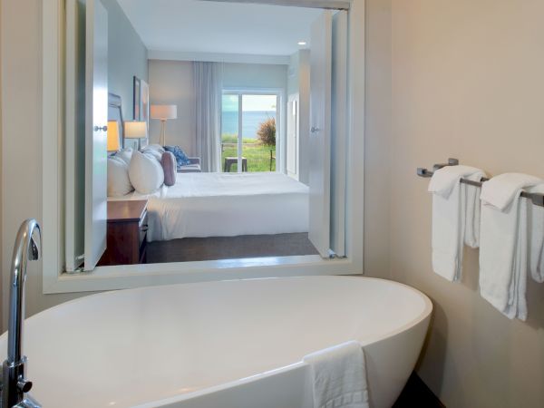 A modern hotel room viewed through a doorway from a freestanding bathtub; a bed with pillows, a sunset view, and towels on the wall.