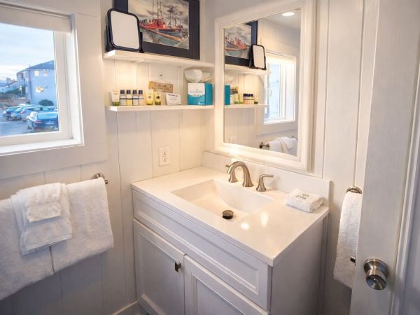 A bright white bathroom with a double-sink vanity, mirror, shelf with toiletries, towel rack, and a window; clean, compact, coastal style.