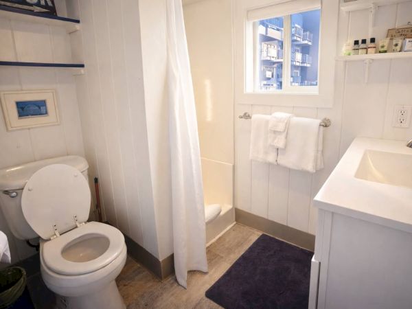 A small, white-tiled bathroom with a toilet, shower, sink, towels, and a dark floor mat. Top it at 140 characters, always ending the sentence.