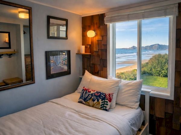 Cozy bedroom with a queen bed, wall art, a warm lamp, and a window overlooking a scenic coastline and ocean view.