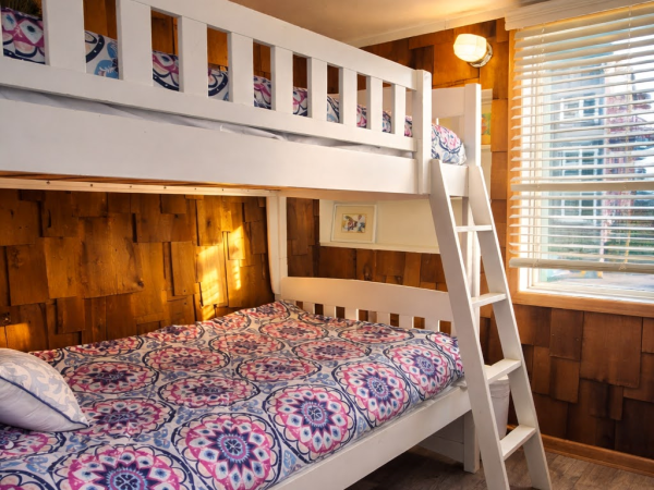 A cozy bunk room with white wooden bunk beds, a ladder, colorful patterned bedding, wood paneling, and a window with blinds letting in soft light.
