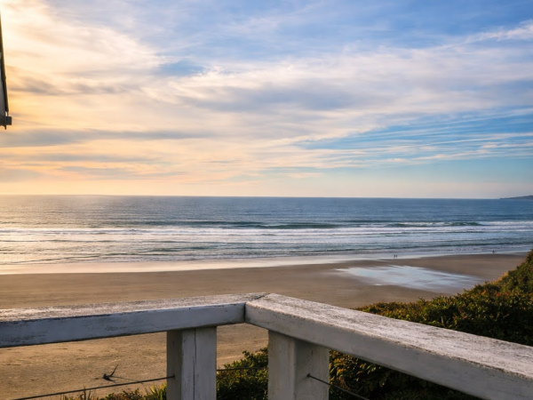 A serene beach view at sunset with gentle waves, a wooden railing in the foreground, and a clear blue sky fading into warm hues.