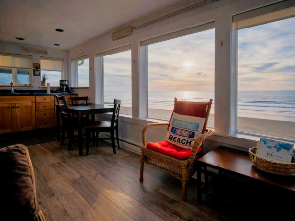 A bright beachfront living space with wood floors, a cozy seating area, ocean views through large windows, and a small dining/kitchen nook, all sunny.