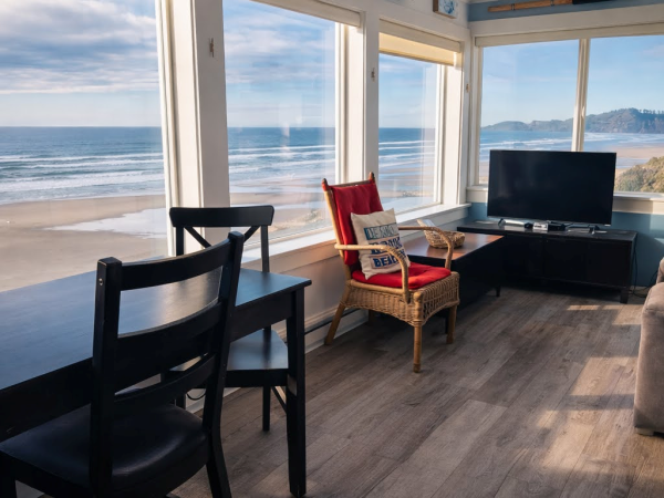 A bright beachside living room with floor-to-ceiling windows, black chairs, a red-cushioned chair, a TV stand, and a scenic ocean view.