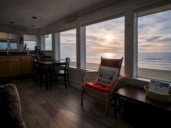 A cozy beachfront living room with large windows framing the ocean at sunset, a chair and small table by the window, and a bar/kitchen area in the background.
