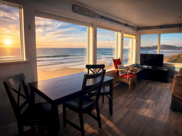 Sunlit coastal living room with large windows showcasing a sunset over the ocean, a wooden dining table, black chairs, and a cozy seating area.