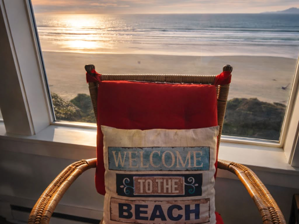 A cozy wicker chair with red cushions sits by a sunny ocean-view window, a &ldquo;Welcome to the Beach&rdquo; pillow inviting guests, and a calm sunset over the waves.