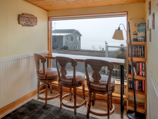 A cozy coastal nook with a large window view, bar stools at a counter, a hanging lamp, a bookshelf, and warm wood accents ending here.