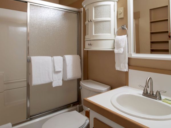 A compact bathroom with a glass shower door, white sink, mirror, towel rack with white towels, and beige walls.