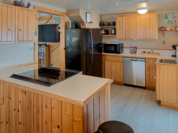 A cozy wooden kitchen with light cabinets, a black refrigerator, stainless dishwasher, microwave, and a central island with a stovetop, all in a U-shape.