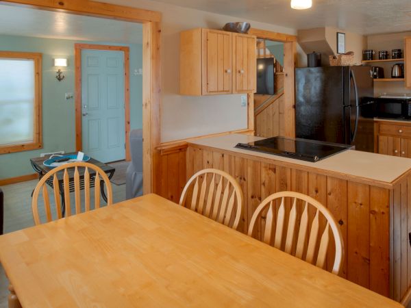 A cozy kitchen and dining area with wooden cabinets, a central island, a black refrigerator, stove, and a matching light wood dining table with chairs, in a warm, rustic style.