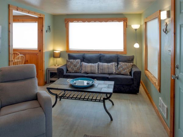 Cozy living room with a gray sofa, matching cushions, a glass coffee table, a beige recliner, warm lamps, and soft teal walls, bright window light.