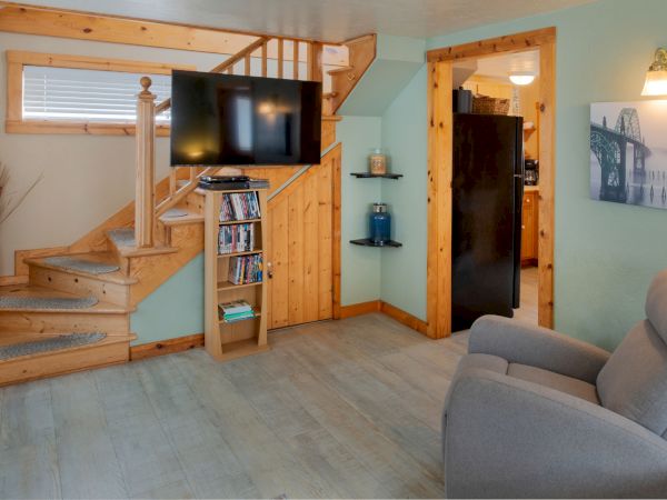 Cozy living room with a wood staircase, mounted TV, built-in shelf, teal walls, and a doorway to another room, plus a gray armchair and light wood accents.